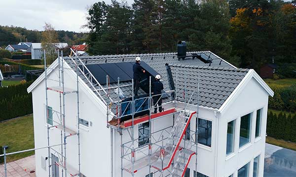 Bostadshus med byggställningar och två installatörer på taket som installerar solceller.