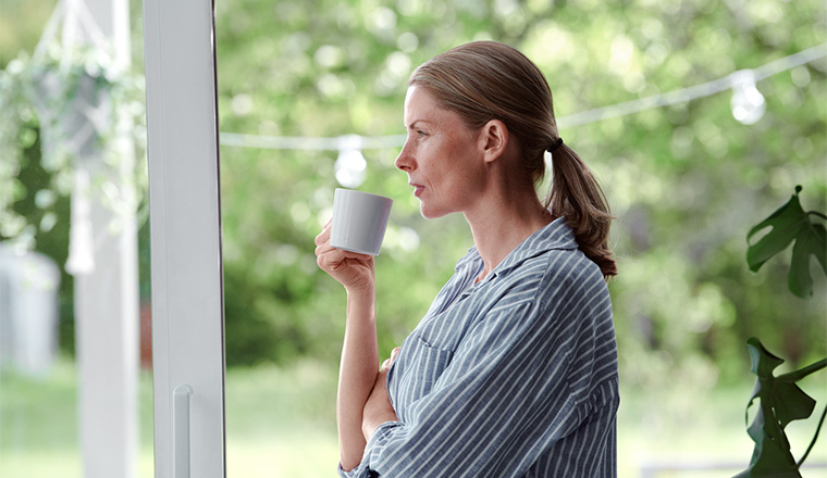 Kvinna med kaffekopp tittar ut mot ett fönster.